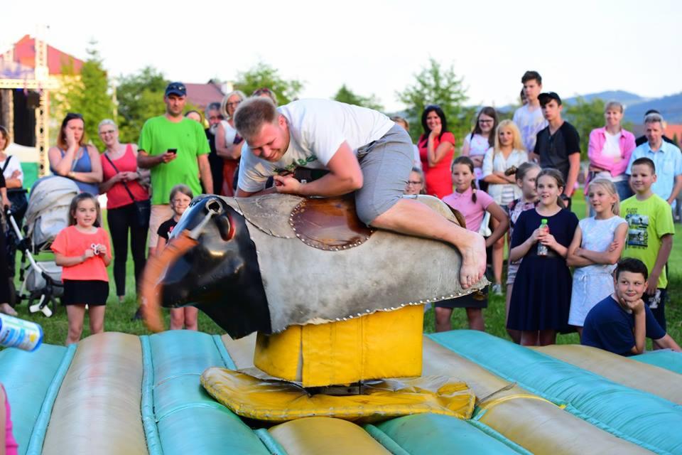 Mężczyzna z grymasem na twarzy próbuje utrzymać się na mechanicznym byku podczas festynu, otoczony grupą obserwujących dzieci i dorosłych.