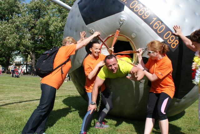 Grupa osób w pomarańczowych koszulkach pomaga mężczyźnie w żółtej koszulce wydostać się z otworu w dużej, szarej kuli z numerem telefonu, na trawniku.