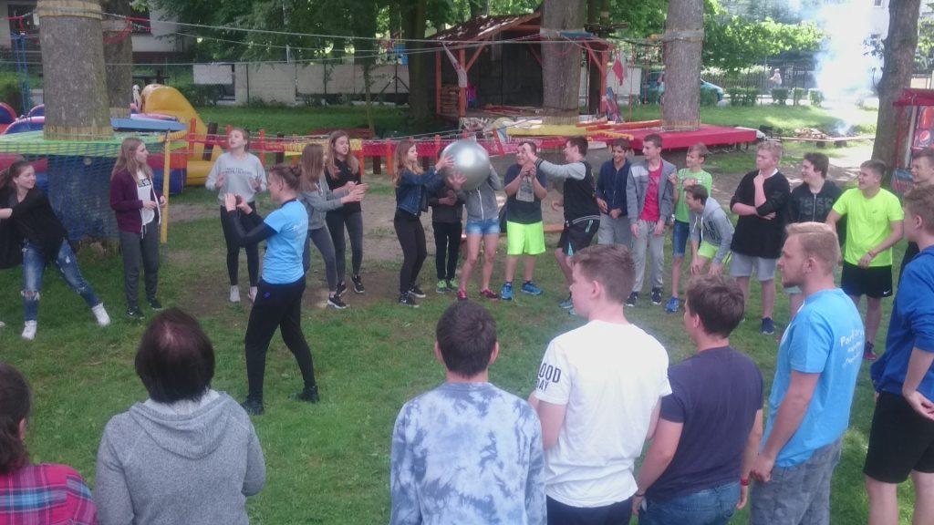 Grupa młodzieży grająca w grę zespołową z dużą srebrną piłką na trawie, w tle plac zabaw linowy i drzewa.