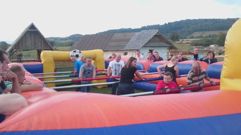 Grupa osób gra w dmuchane piłkarzyki na świeżym powietrzu podczas imprezy integracyjnej, w tle budynki gospodarcze i zalesione wzgórza.