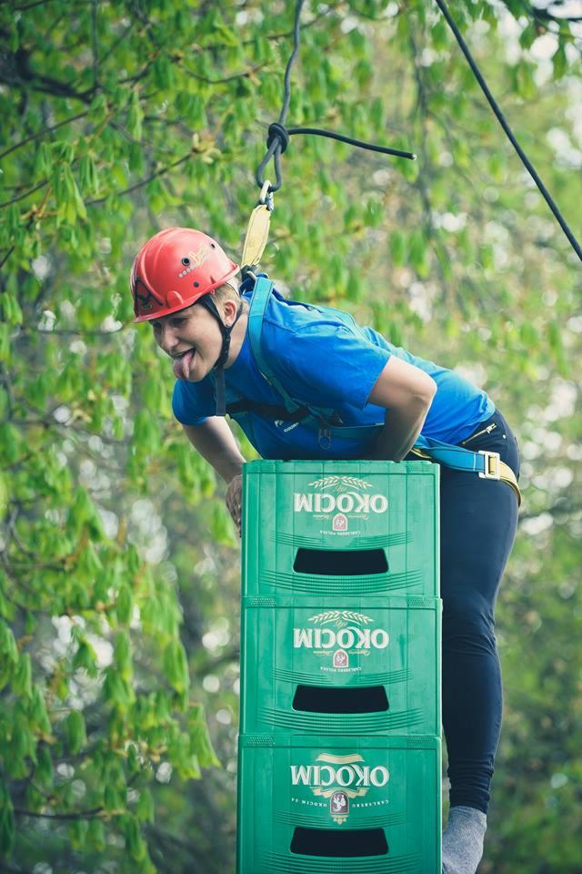 Osoba w kasku i uprzęży, z wyciągniętym językiem, opiera się na zielonych skrzynkach z napisem 'Okocim' podczas zabawy na wysokościowym torze przeszkód w Kudowie-Zdroju.