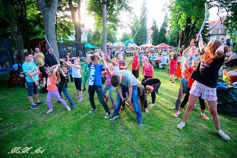 Grupa dzieci uczestniczy w zabawie z niskim limbo na trawie podczas imprezy plenerowej, w tle widoczni dorośli, trampolina i budynki.
