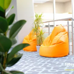 Jasne, nowoczesne wnętrze z dwoma pomarańczowymi pufami typu bean bag, rośliną w doniczce i fragmentem schodów z metalową barierką na wzorzystej podłodze.