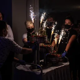 Trzy poziomowy tort udekorowany owocami i czekoladą, z płonącymi fontannami, niesiony przez kelnerki w maskach i rękawiczkach.