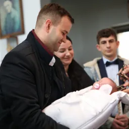 Uśmiechnięty mężczyzna trzyma zawinięte w biały kocyk niemowlę z różowym smoczkiem podczas chrztu, w tle kobieta i mężczyzna w muszce.