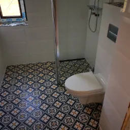 Wykończona łazienka z białą armaturą, kabiną prysznicową i wzorzystą, ciemną podłogą w stylu retro, widoczne białe płytki na ścianach.