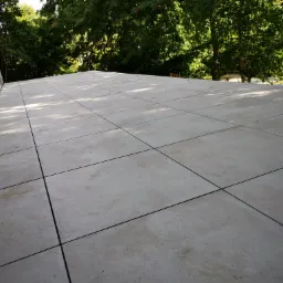 Ułożona, jasnoszara posadzka z płytek na zewnątrz budynku, z widocznym fragmentem elewacji i zielenią drzew w tle.