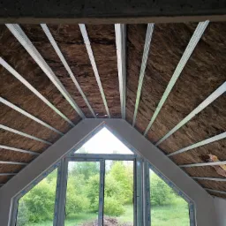 Poddasze z oknem dachowym w trakcie ocieplania, widoczne metalowe profile konstrukcyjne i wełna mineralna, na zewnątrz zielony krajobraz.