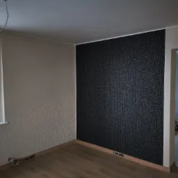 Pokój po remoncie z częściowo wykończonymi ścianami: jedna ściana w ciemnej tapecie w geometryczne wzory, druga w jasnej tapecie z fakturą, drewniana podłoga, listwy przypodłogowe z zamontowanymi...