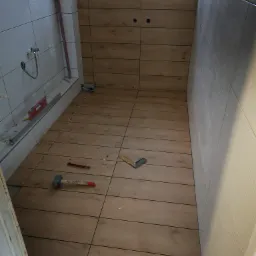 Łazienka w trakcie remontu: brązowe płytki imitujące drewno ułożone na podłodze i ścianie, białe płytki z falistą strukturą na pozostałych ścianach, widoczne narzędzia glazurnicze, poziomica...