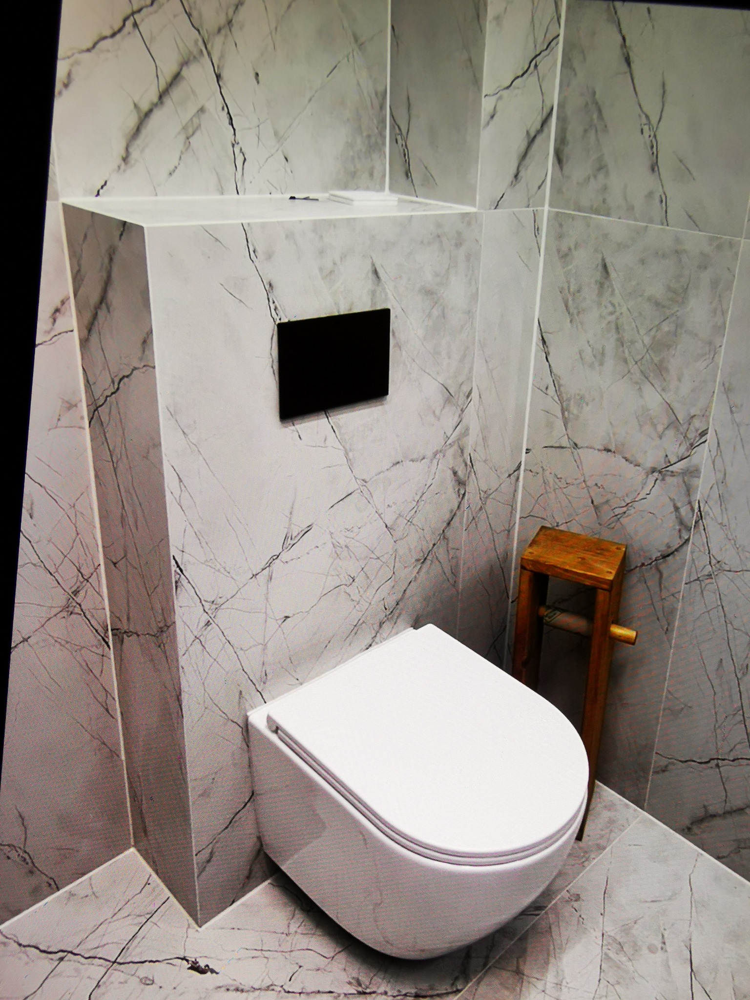 Nowoczesna toaleta podwieszana w łazience wykończonej płytkami imitującymi marmur. Drewniany stołek obok. Minimalistyczny design, eleganckie wykończenie.