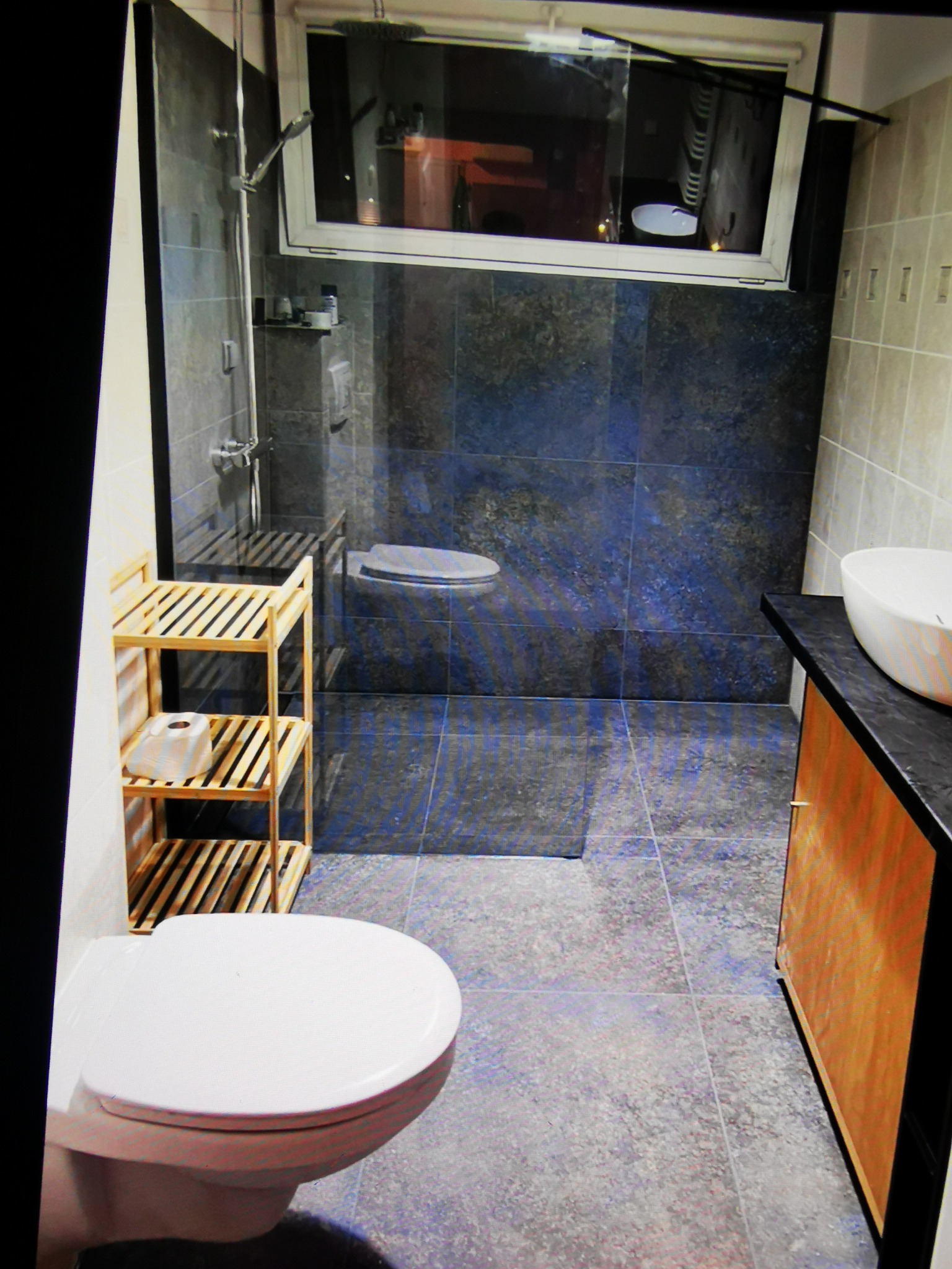 Nowoczesna łazienka z szarymi płytkami, kabiną prysznicową typu walk-in, drewnianą szafką pod umywalką i toaletą w tle. Minimalistyczny design z akcentami drewna.