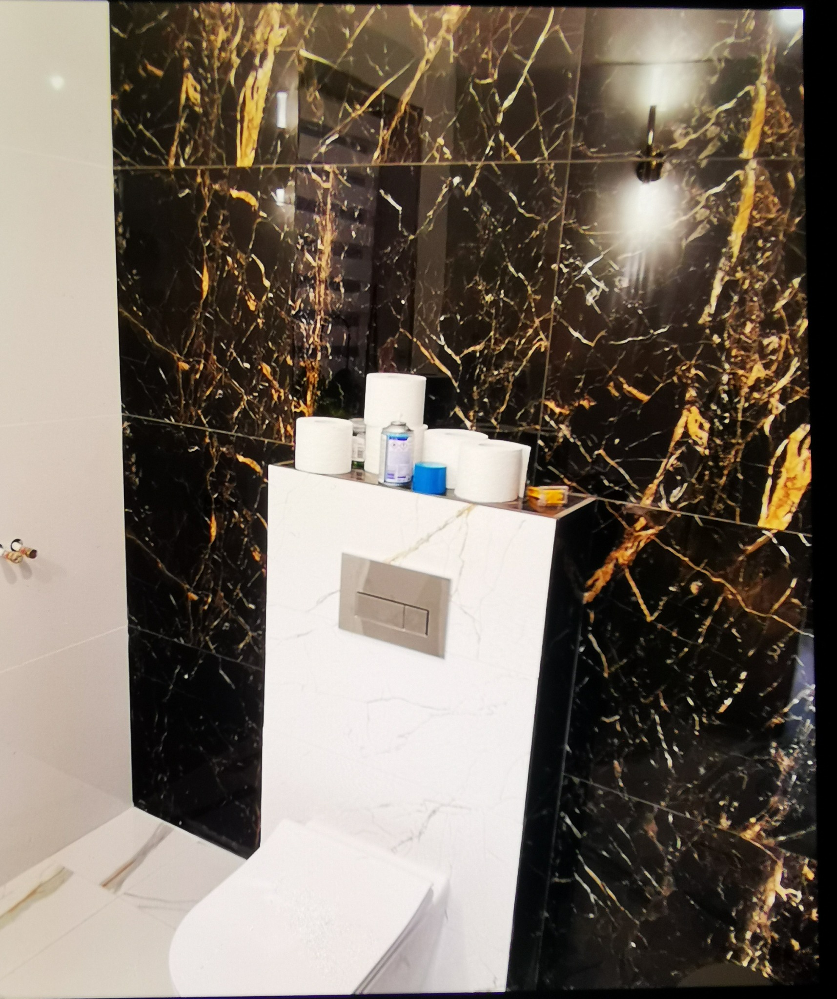 Wykończona łazienka z czarno-złotymi płytkami imitującymi marmur, białą zabudową toalety i akcesoriami. Widoczne detale wykończenia i armatura.