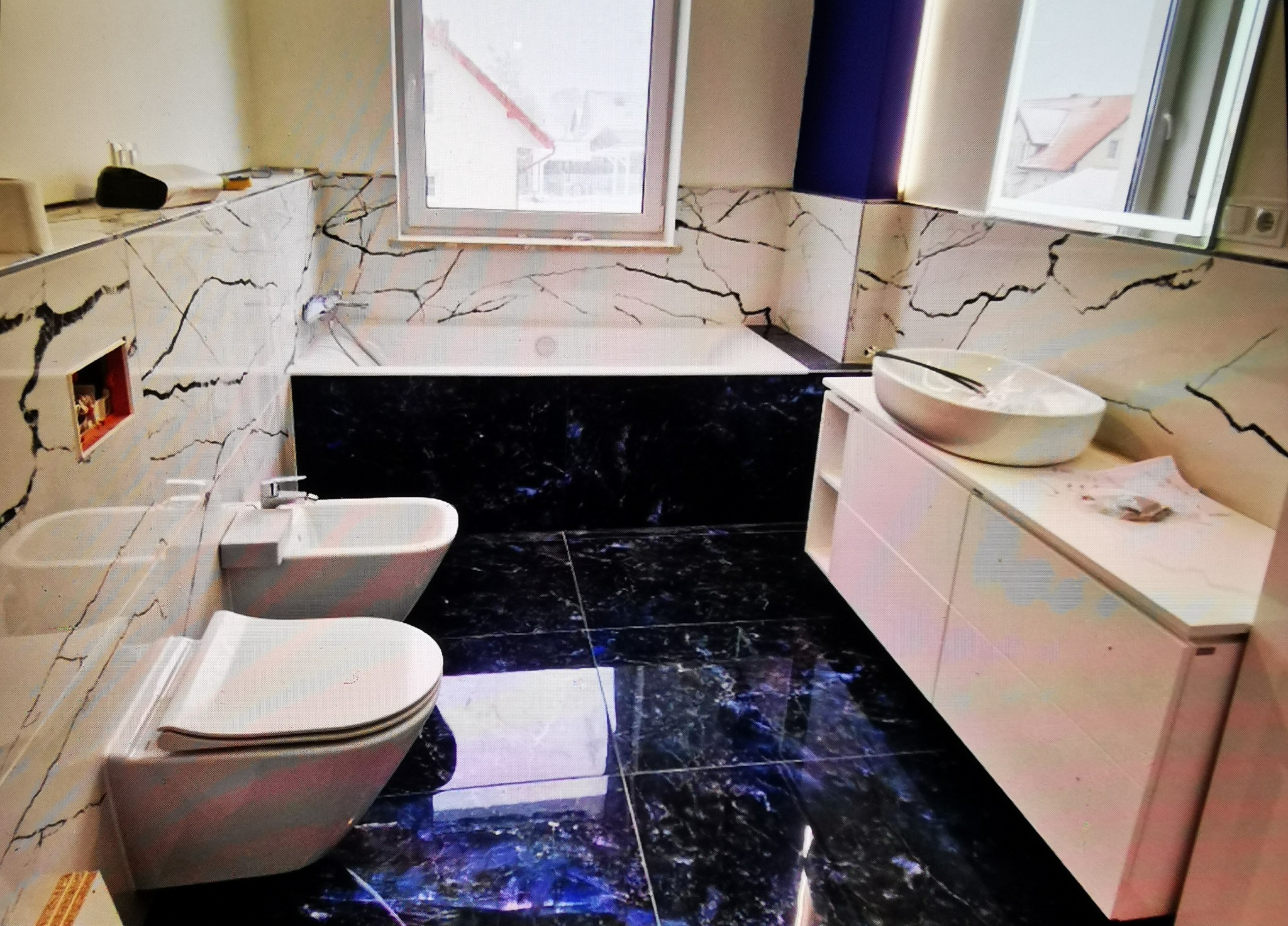 Wykończona łazienka z czarnymi płytkami imitującymi marmur na podłodze i ścianach, białą armaturą i szafką pod umywalką. Widoczne okno i lustro z podświetleniem.