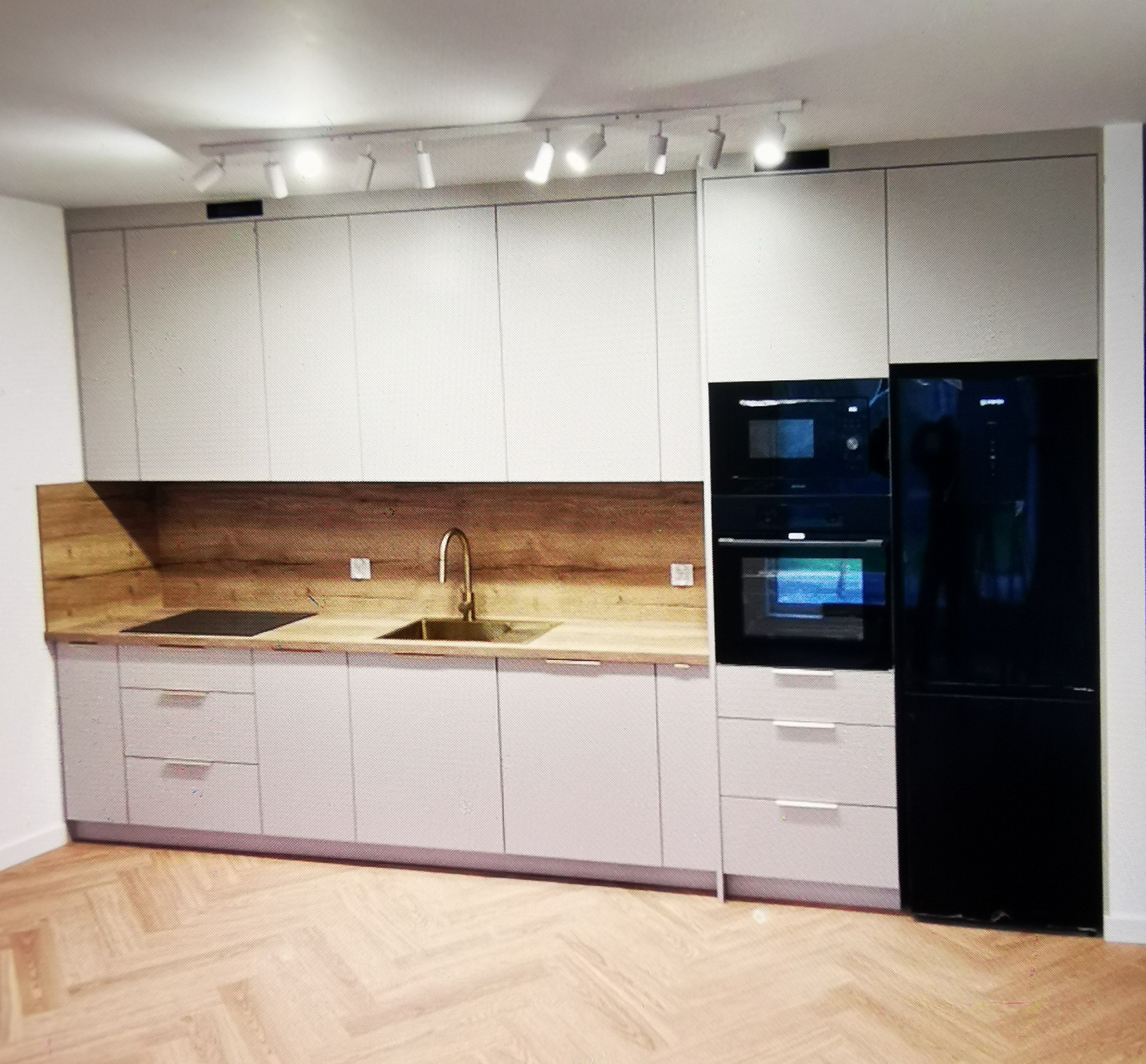 Nowoczesna kuchnia z białymi szafkami, drewnianym blatem i czarnym sprzętem AGD. Uwagę przykuwa złota bateria umywalkowa i parkiet ułożony w jodełkę.