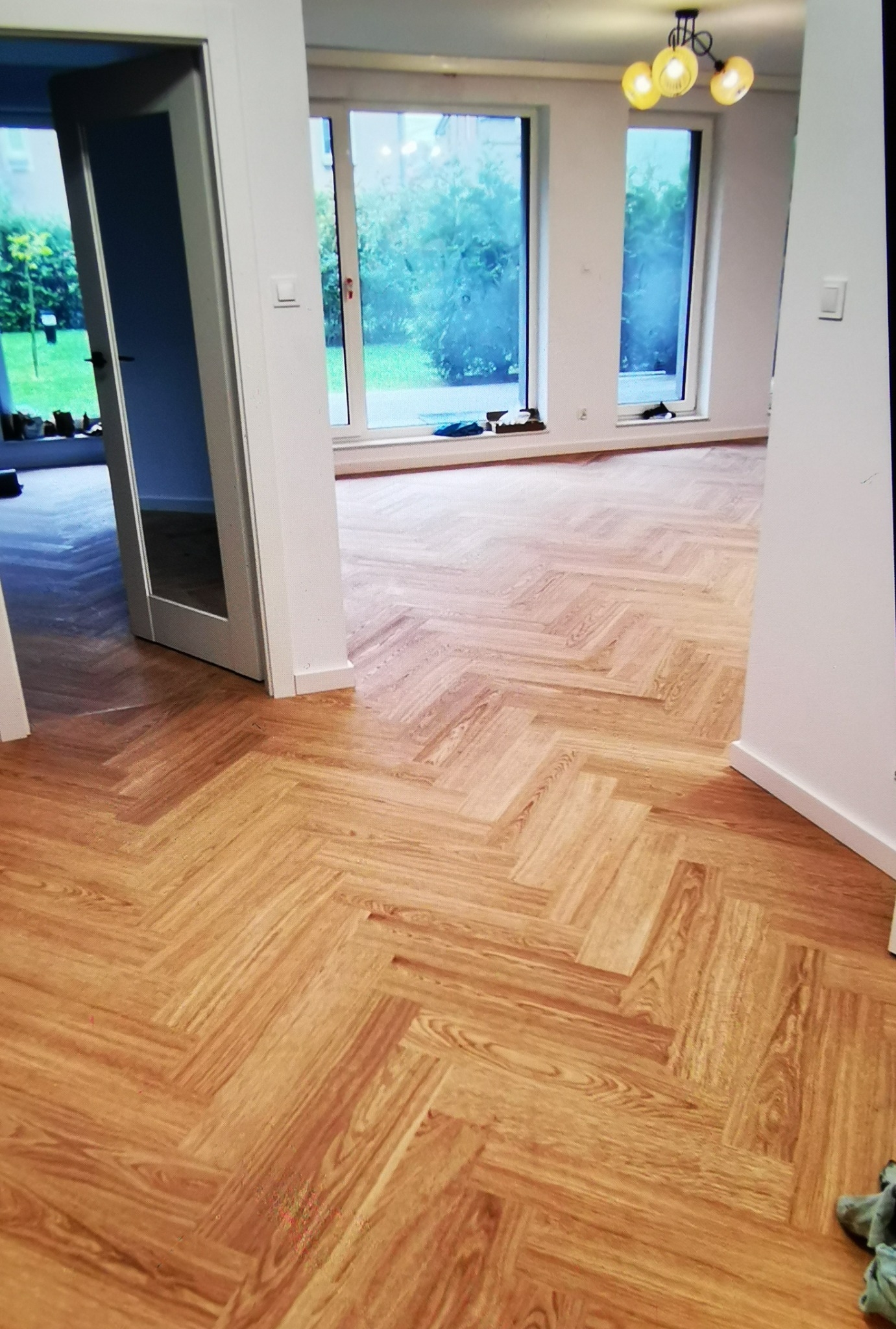 Parkiet ułożony w jodełkę francuską w jasnym wnętrzu z oknami i otwartymi drzwiami. Widoczny fragment sufitu z lampą. Naturalne światło dzienne.