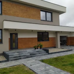 Nowoczesny dom jednorodzinny z drewnianą elewacją, szarymi schodami wejściowymi i podjazdem z kostki brukowej, oświetlony lampami zewnętrznymi, z widocznym wnętrzem garażu i zielenią w donicach.