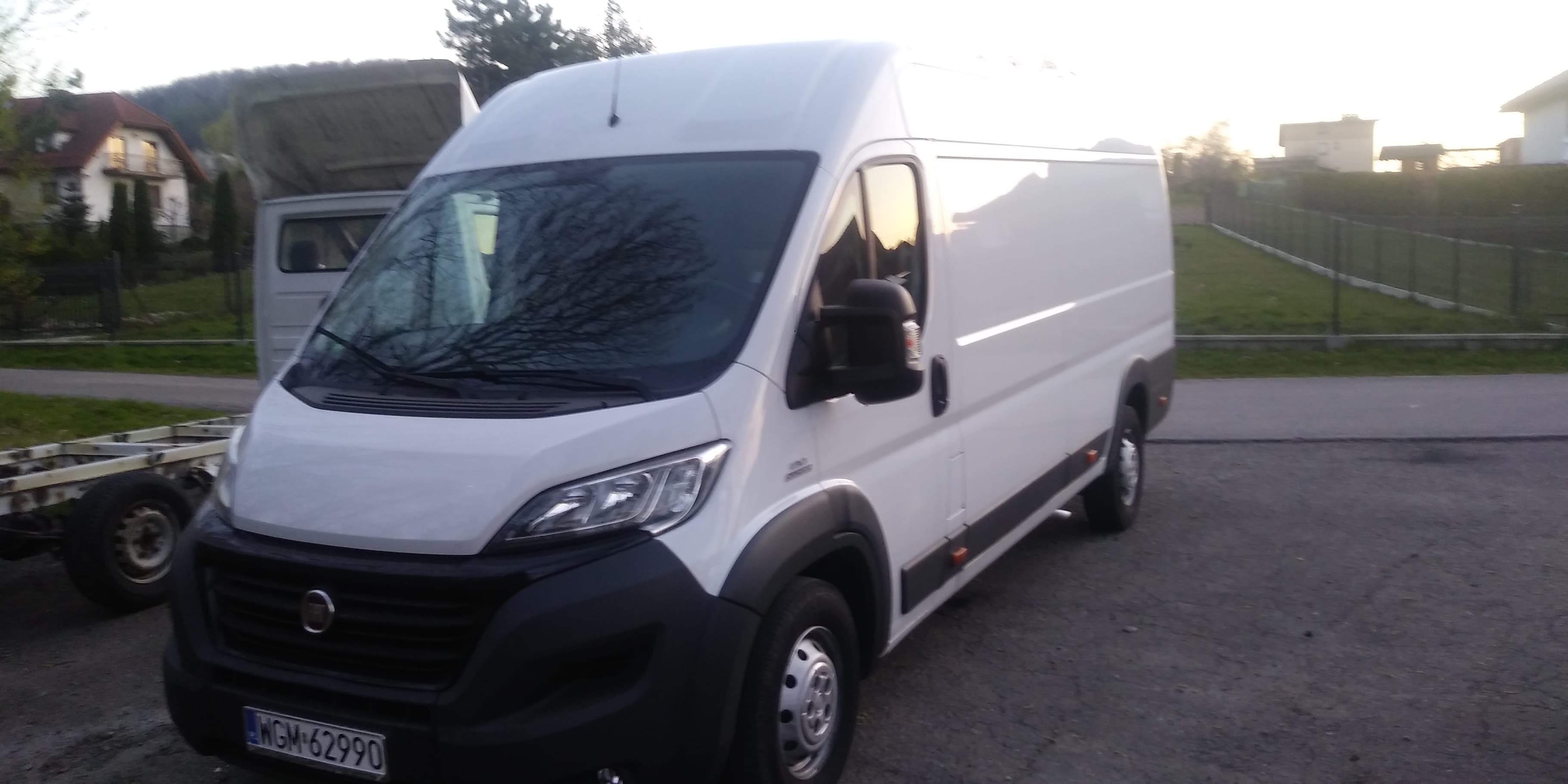 Biały bus dostawczy Fiat Ducato na parkingu, z widocznym numerem rejestracyjnym WGM-62990, w tle częściowo widoczna przyczepa i zabudowa domów jednorodzinnych.