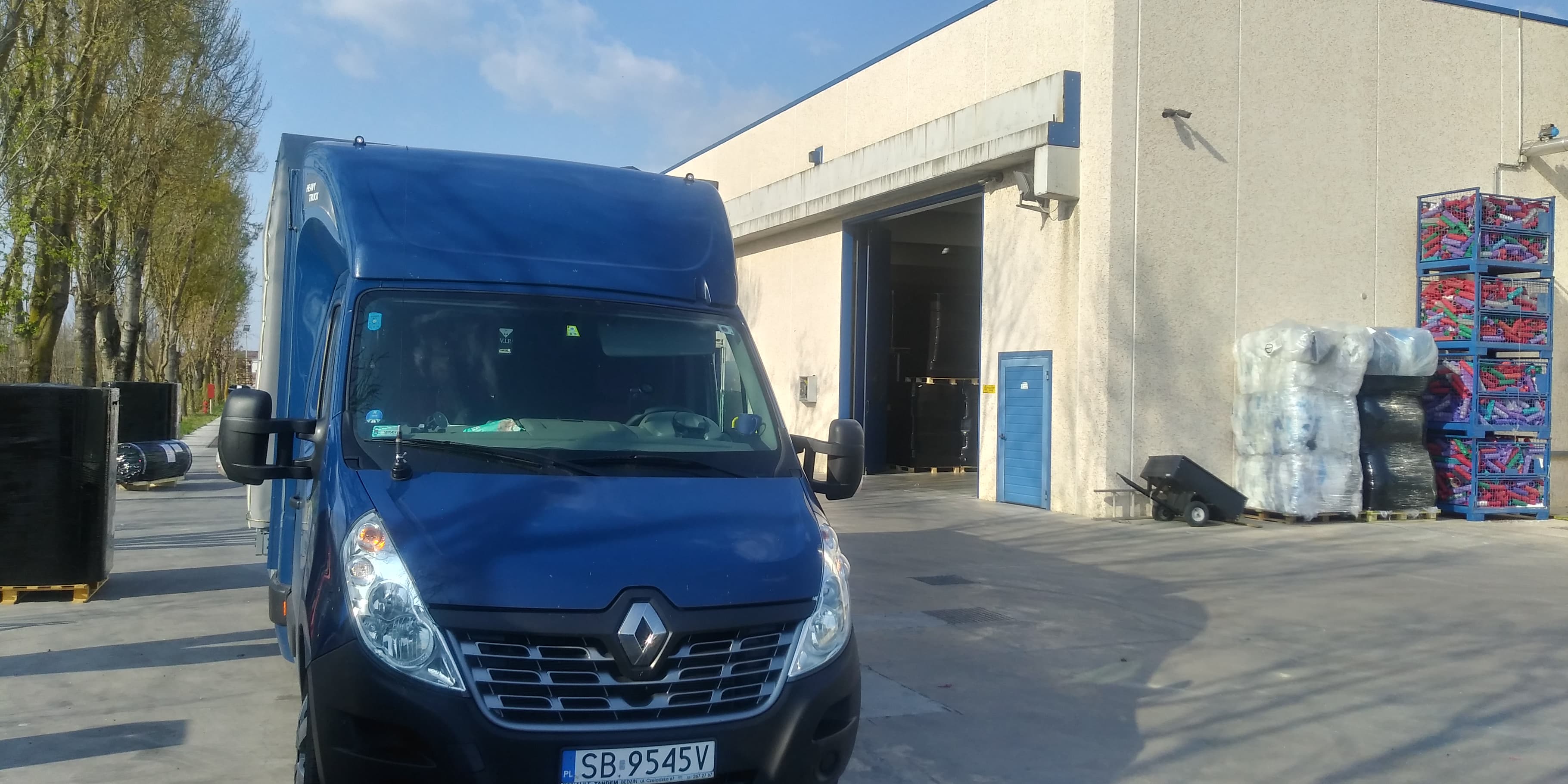 Niebieski bus Renault Master zaparkowany przed magazynem z otwartą bramą, w tle palety z towarem, po prawej stronie regały z kolorowymi produktami w koszach.