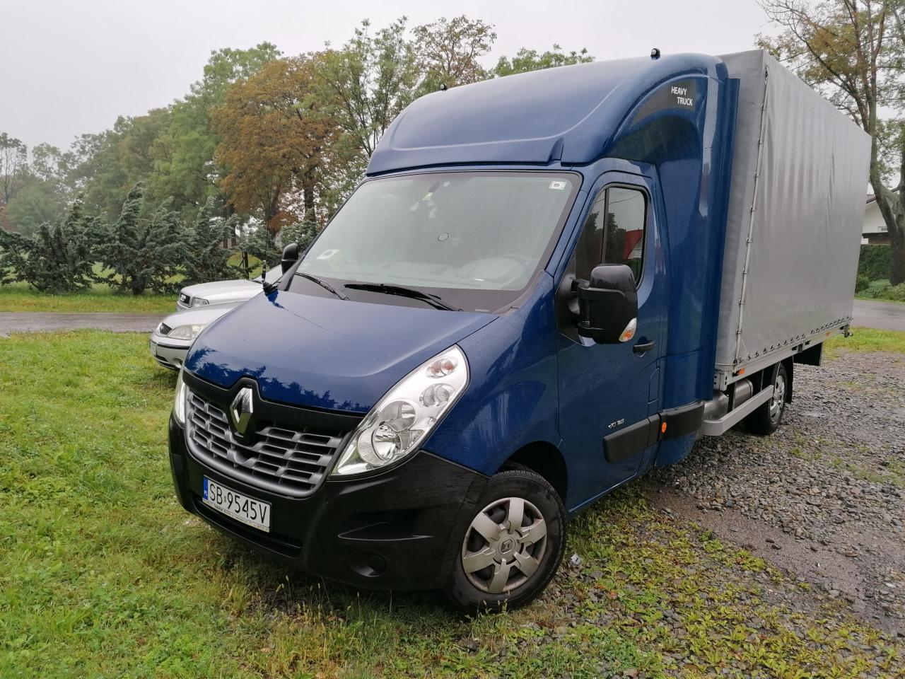 Niebieski samochód dostawczy Renault Master z plandeką, zaparkowany na trawie w pochmurny dzień, widoczny numer rejestracyjny i napis 'Heavy Truck' na kabinie.