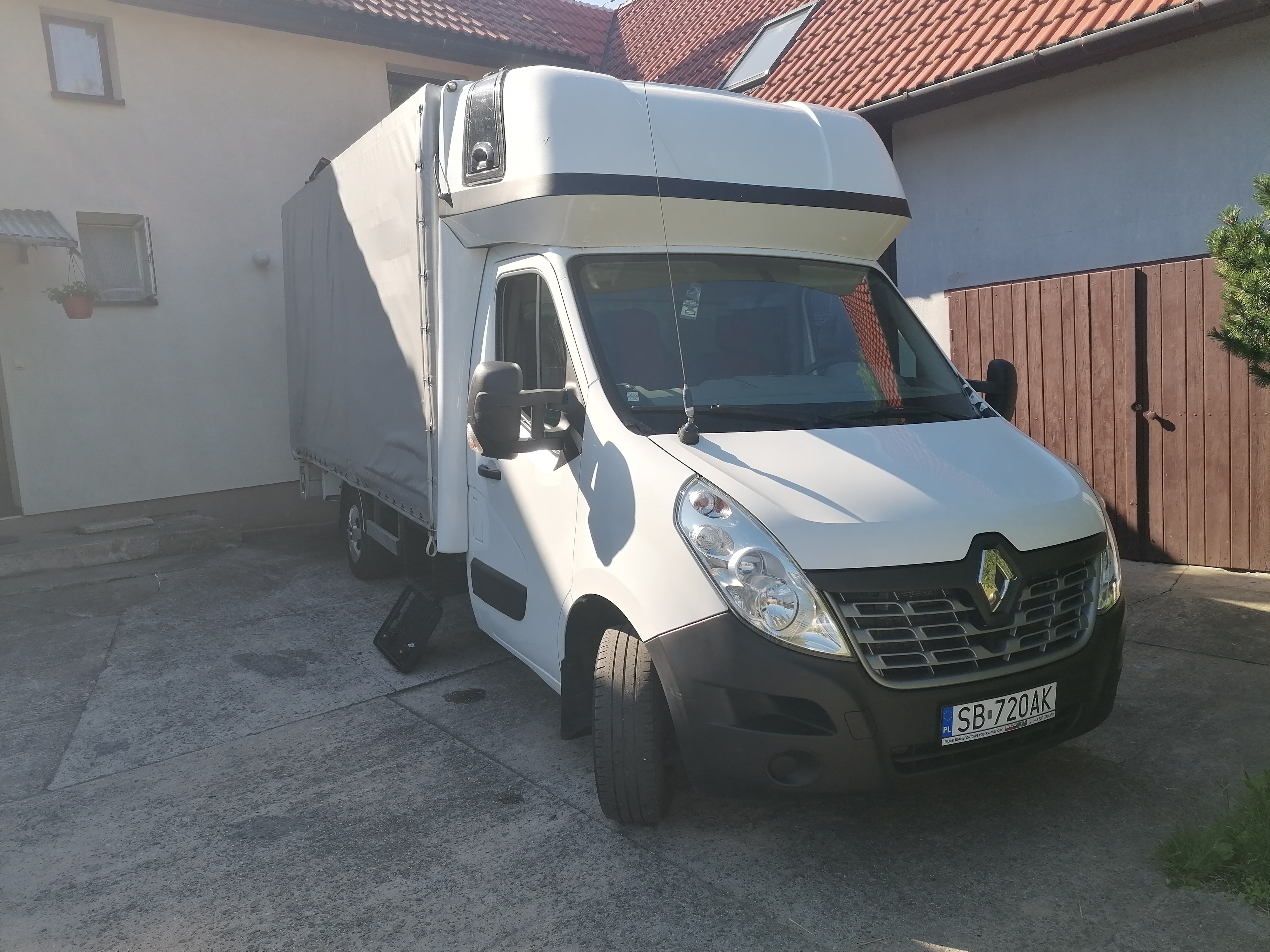 Biały samochód dostawczy Renault Master z plandeką na betonowym podjeździe przed domem z czerwoną dachówką; obok leży czarna skrzynka.