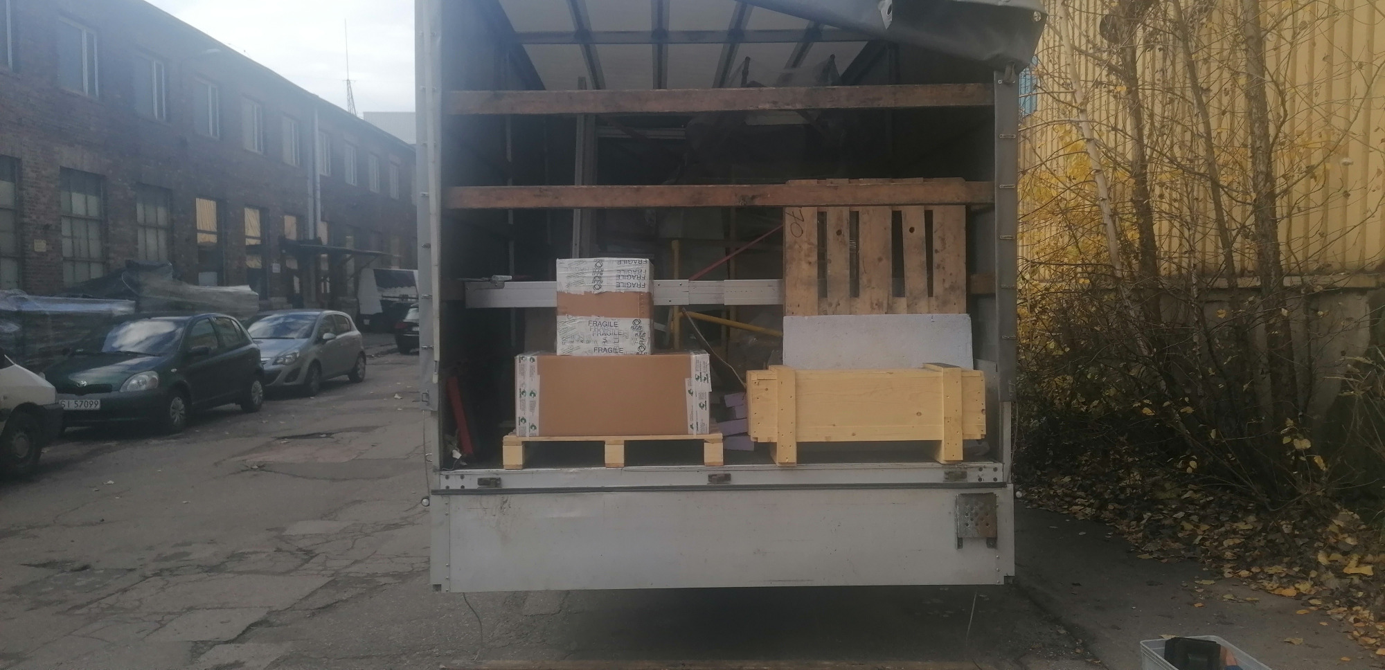 Wnętrze przestrzeni ładunkowej samochodu dostawczego wypełnione kartonami i drewnianymi skrzyniami, zabezpieczonymi na paletach, widok z zewnątrz na ulicę z zaparkowanymi autami i budynkiem...