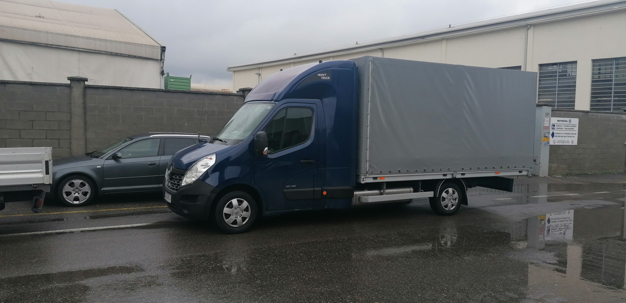 Niebieski samochód dostawczy z plandeką zaparkowany na mokrym placu przemysłowym, obok szarego samochodu osobowego i szarego muru.