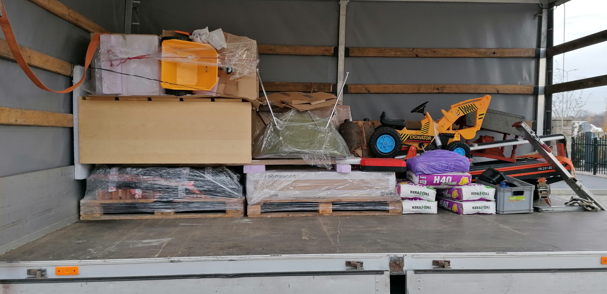 Wnętrze przestrzeni ładunkowej z różnorodnymi przedmiotami zabezpieczonymi do transportu, w tym palety z materiałami budowlanymi, zabawka - koparka, meble i inne przedmioty.