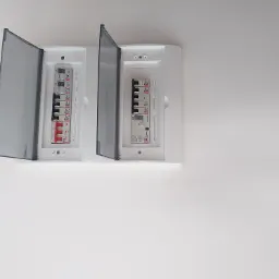 Dwie białe skrzynki elektryczne z otwartymi, przydymionymi, plastikowymi osłonami na białej ścianie, widoczne bezpieczniki i oznaczenia obwodów.