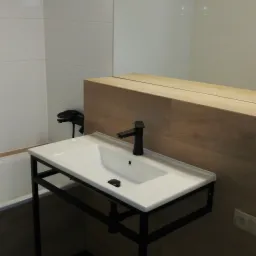 Nowoczesna umywalka nablatowa z czarną armaturą na metalowym stelażu w łazience wykończonej białymi płytkami i panelami drewnopodobnymi.