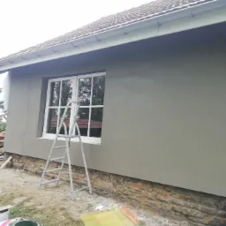 Świeżo otynkowana ściana domu z widoczną drabiną przy oknie, fragmentem kamiennej podmurówki i stertą drewna w tle.