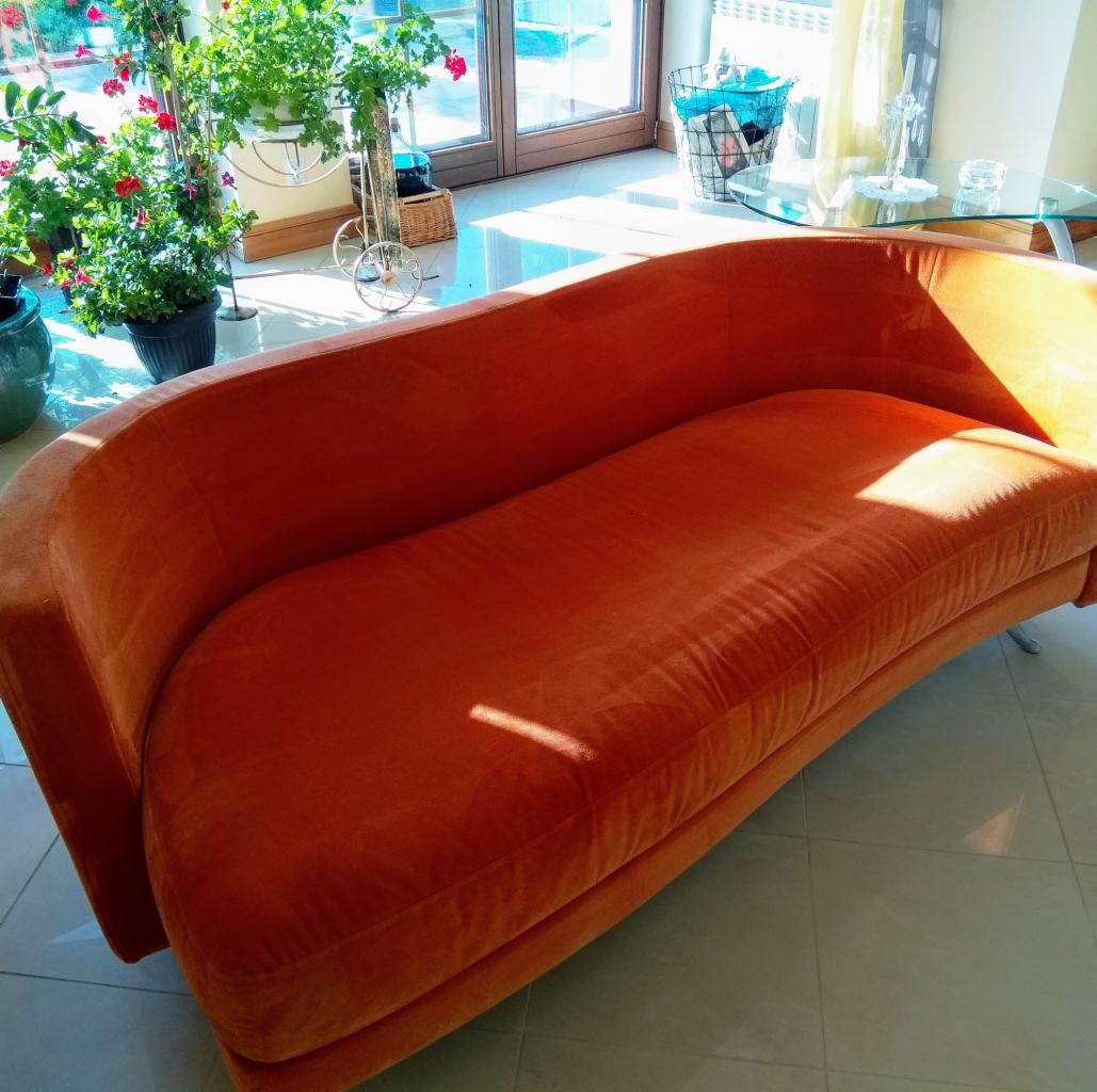 Nowoczesna, pomarańczowa sofa z aksamitną tapicerką stoi na jasnych płytkach podłogowych, obok okna z widokiem na rośliny doniczkowe i dekoracyjny wózek.