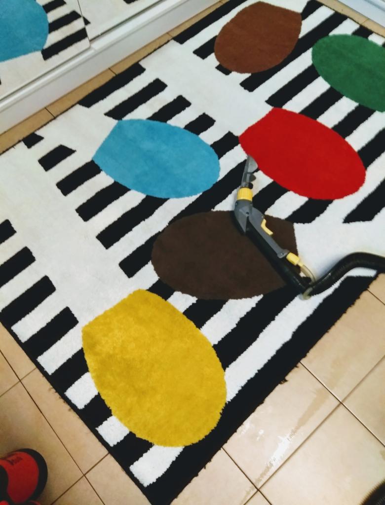 Czyszczenie wzorzystego dywanu z kolorowymi plamami przy użyciu odkurzacza piorącego z dyszą, widoczne ślady wilgoci na płytkach podłogowych.