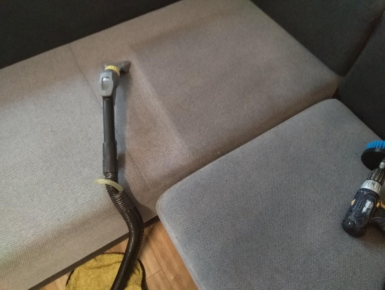 Szara sofa podczas czyszczenia: widoczna dysza odkurzacza piorącego na siedzisku oraz wiertarka z szczotką do czyszczenia tapicerki na poduszce. Podłoga częściowo widoczna, z żółtą ścierką.
