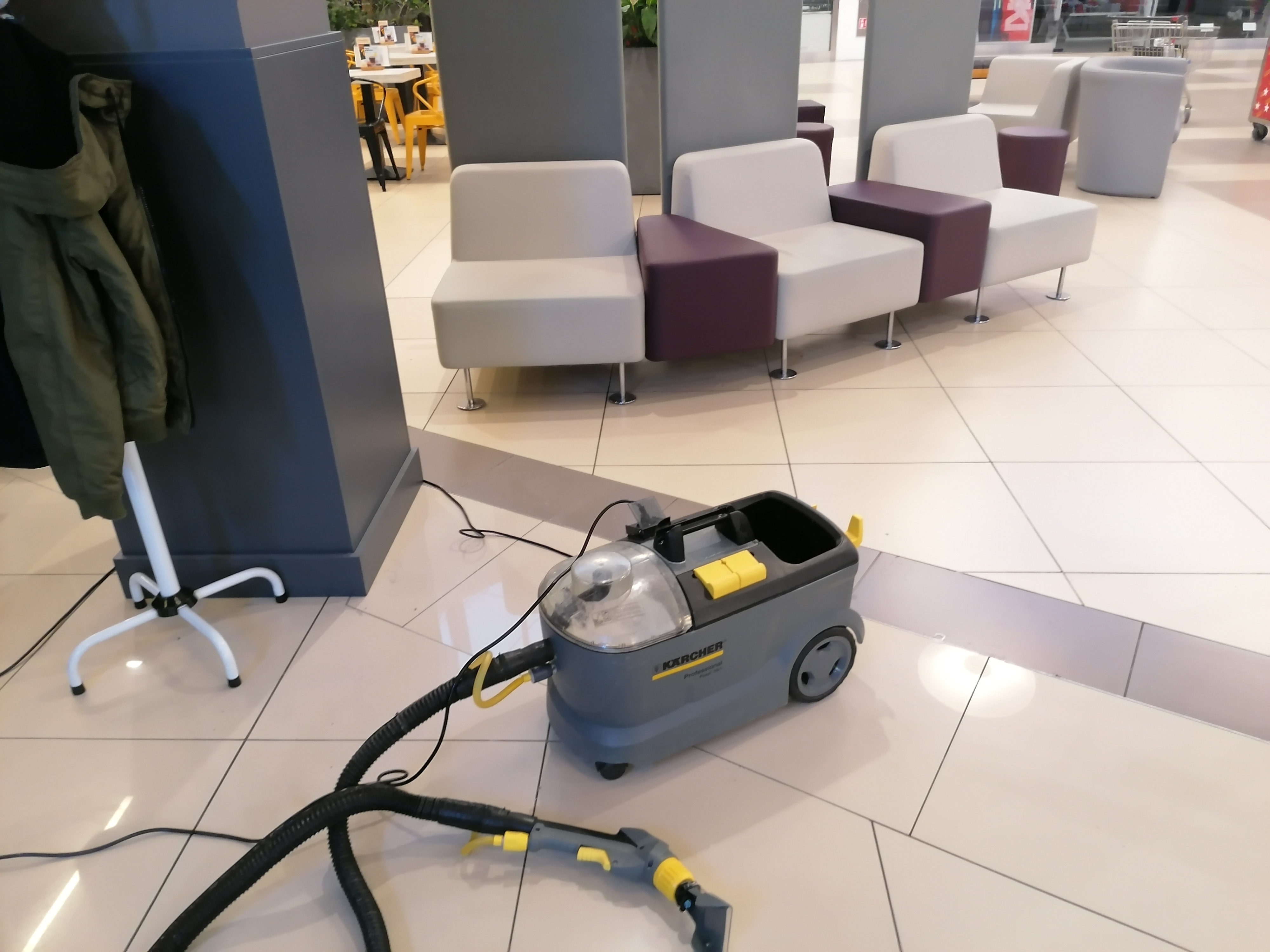 Profesjonalny odkurzacz piorący Karcher na jasnej, kafelkowej podłodze w przestrzeni publicznej, prawdopodobnie biurowej lub handlowej, z widocznymi w tle nowoczesnymi sofami i wieszakiem na ubrania.