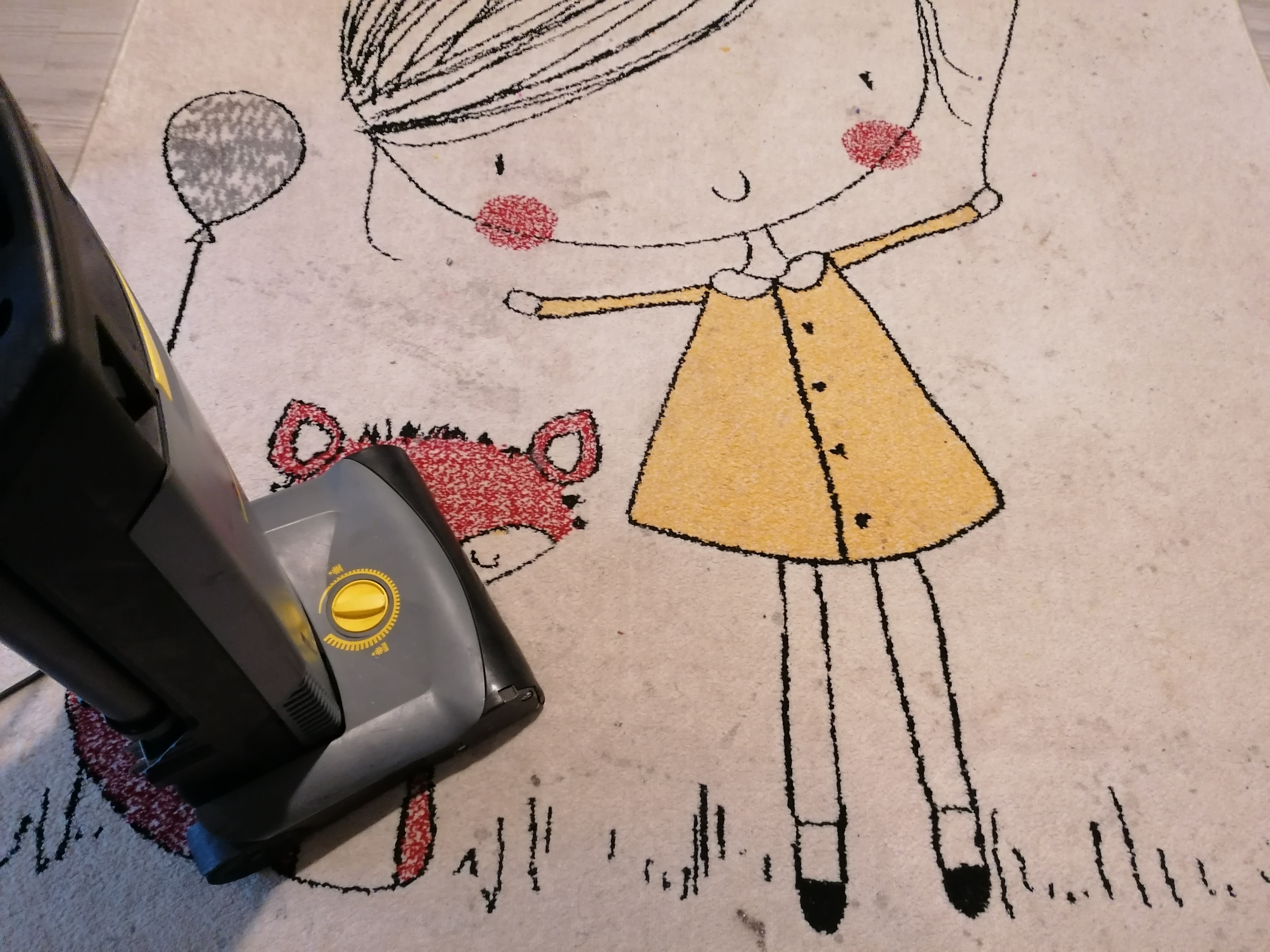 Szary odkurzacz piorący na dywanie z dziecięcym wzorem dziewczynki z balonem i liskiem, widoczne zabrudzenia przed czyszczeniem.
