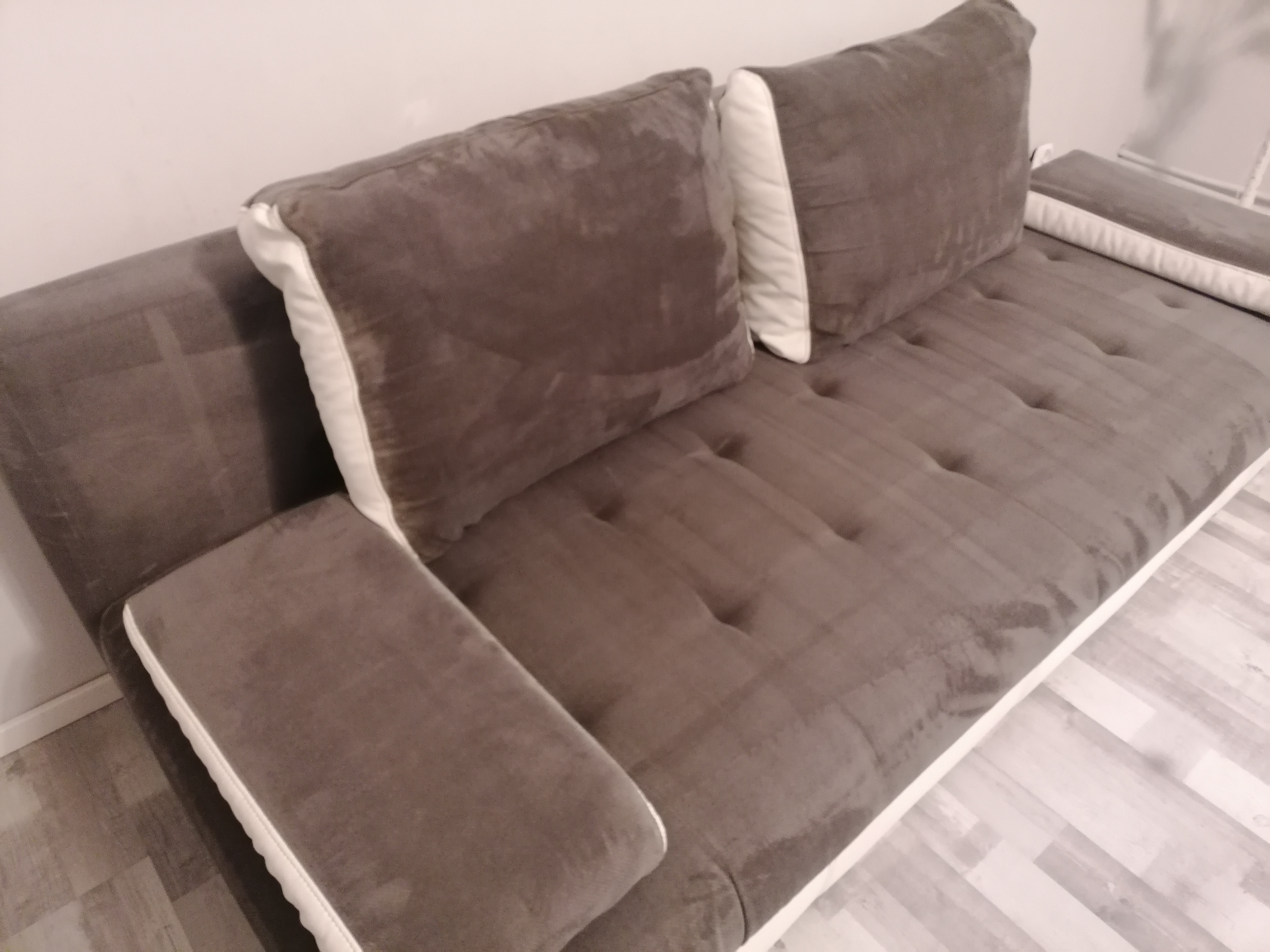 Szara, rozkładana kanapa z welurową tapicerką i białymi lamówkami na poduszkach i oparciach, widoczna z góry, na podłodze z paneli.