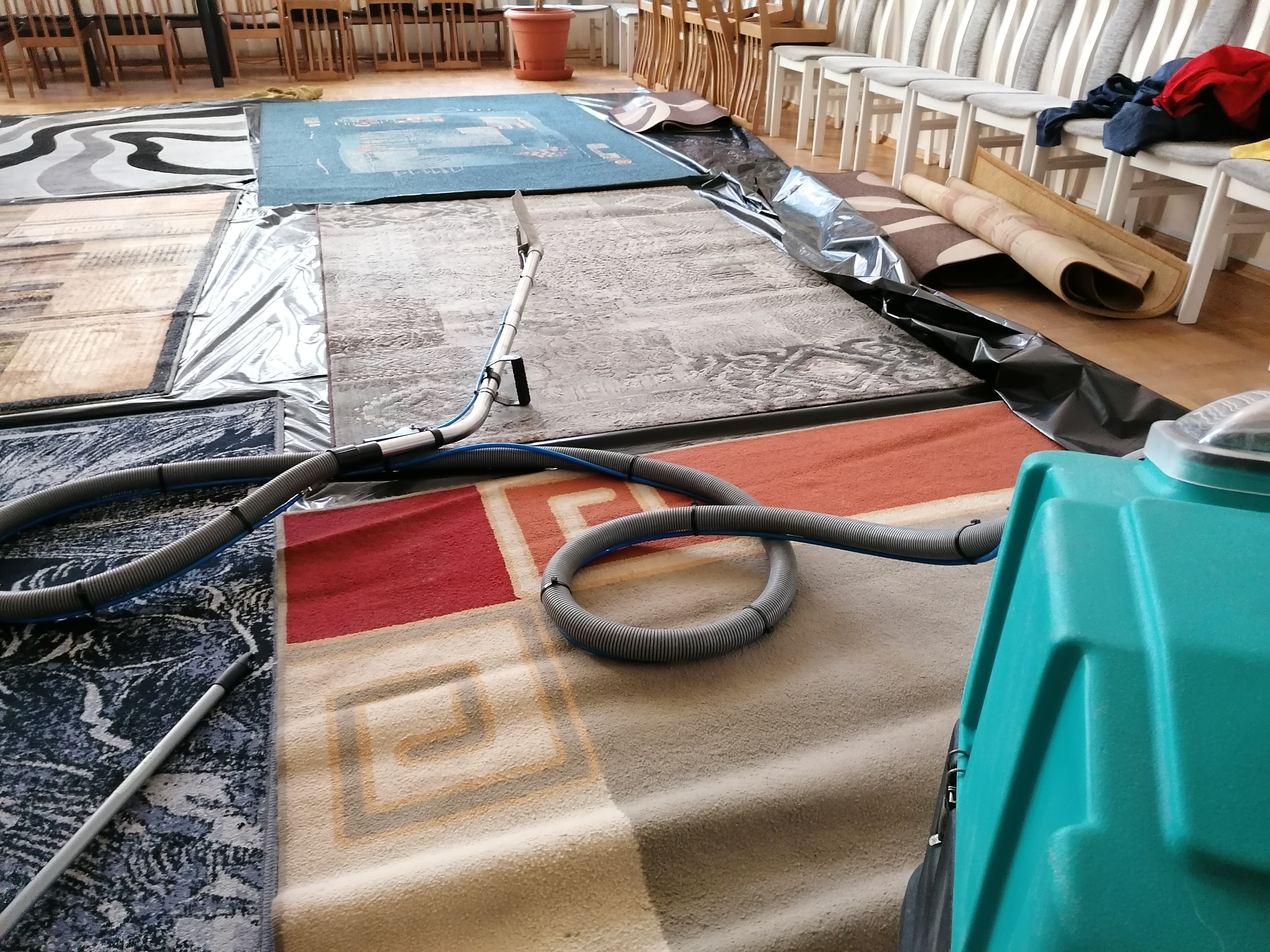 Maszyna do czyszczenia dywanów z wężem i lancą na tle rozłożonych dywanów o różnych wzorach i kolorach, zabezpieczonych folią. W tle widoczne krzesła i inne dywany zwinięte w rulony.