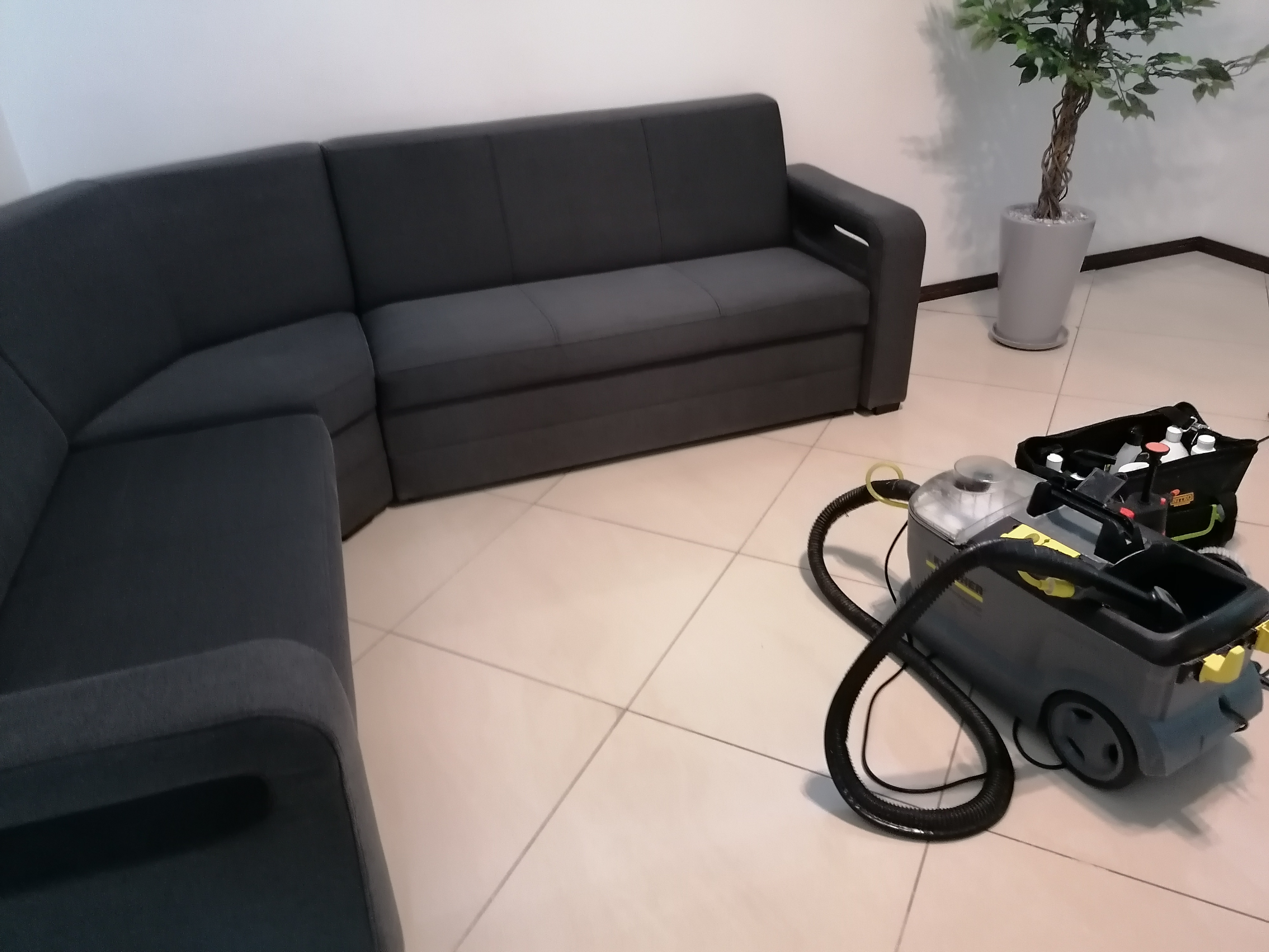 Szara, narożna kanapa z tapicerką materiałową i profesjonalny odkurzacz piorący Kärcher z akcesoriami na kafelkach w salonie.