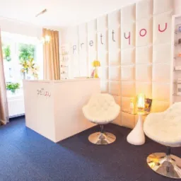 Jasne, przestronne wnętrze salonu z recepcją, dwoma pikowanymi fotelami na chromowanych podstawach, szklanym stolikiem i podświetlaną ścianą z logo firmy.