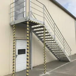 Zewnętrzne, stalowe schody z balustradą, prowadzące do drzwi na podwyższeniu, z elementami zabezpieczającymi w postaci czarno-żółtych pasów na podporach.