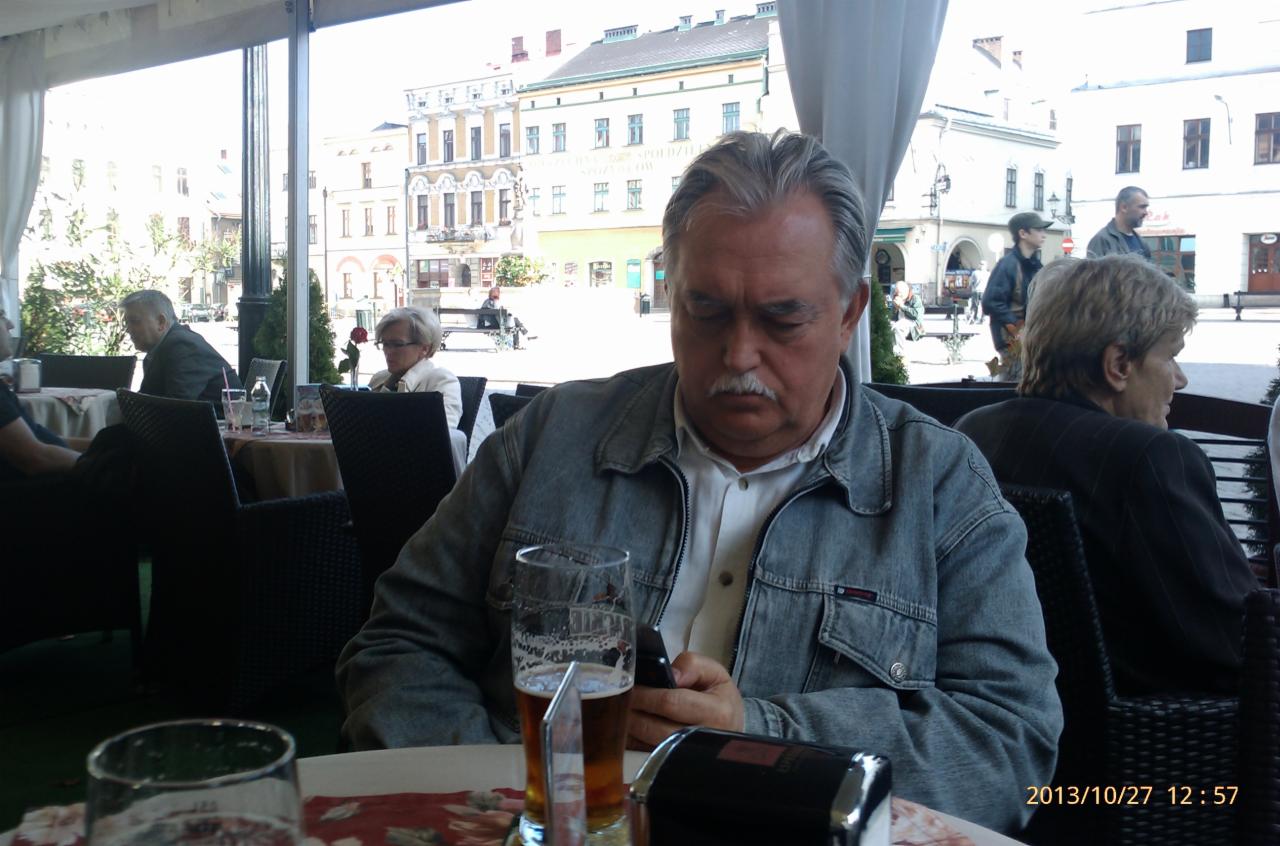 Mężczyzna w dżinsowej kurtce siedzi przy stoliku na zewnątrz kawiarni, przeglądając telefon. Na stole stoi szklanka z piwem i popielniczka. W tle budynki miejskie.