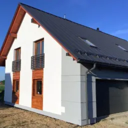 Nowoczesny dom jednorodzinny z ciemnoszarym dachem blaszanym i białymi ścianami, widoczne okna z drewnianym wykończeniem i mini balkonami, szara część garażowa z rynnami.