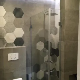 Nowoczesna łazienka z kabiną prysznicową ze szklanymi drzwiami, heksagonalne płytki w odcieniach szarości i bieli na ścianie, minimalistyczna toaleta podwieszana i podświetlenie LED podsufitowe.