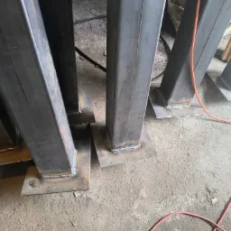 Metalowe słupy o kwadratowym przekroju, z przyspawanymi podstawami, ustawione w rzędzie na betonowej posadzce, widoczne ślady spawania.