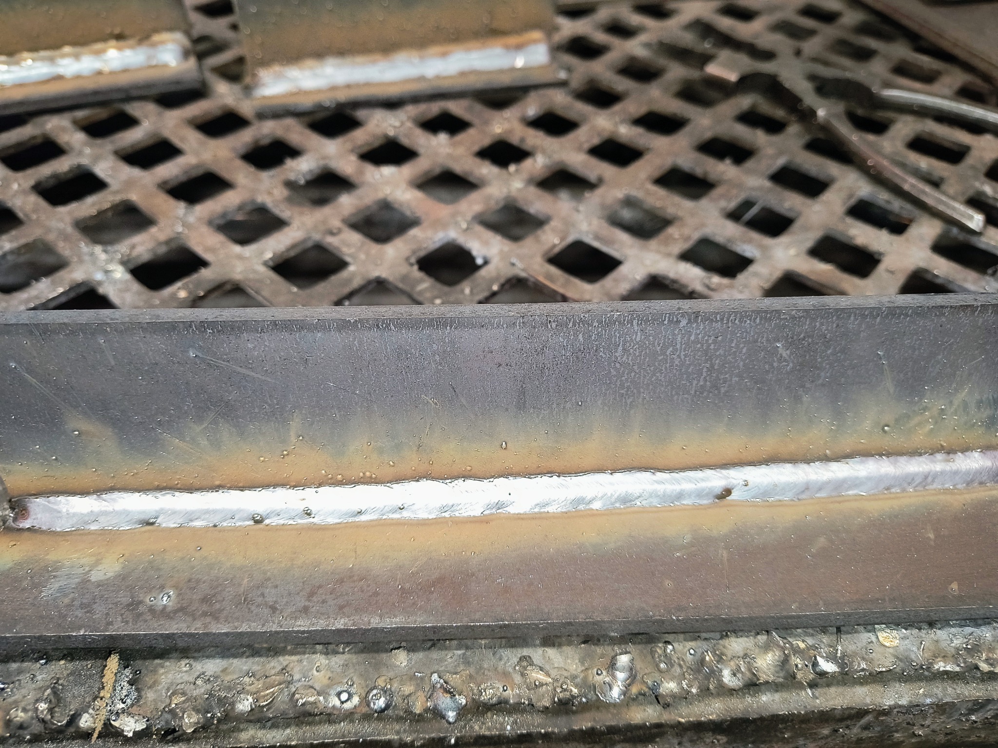 Spoina na metalowym profilu kątowym, widoczna delikatna strefa wpływu ciepła, na tle kratownicy pomostowej.