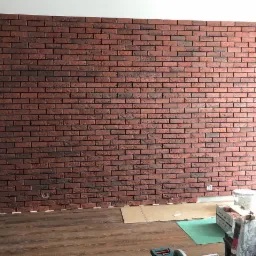 Ściana wykończona czerwoną cegłą klinkierową, widoczne narzędzia i materiały budowlane na podłodze z paneli, dolny fragment ściany jeszcze nie wykończony.
