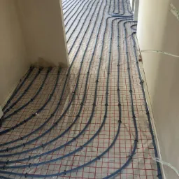 Niebieskie rury ogrzewania podłogowego ułożone na siatce z czerwonymi liniami w surowym wnętrzu przed wylaniem posadzki.