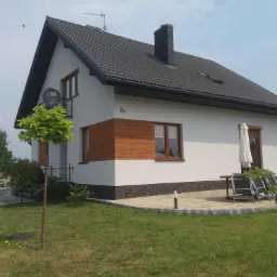 Nowoczesny dom jednorodzinny z ciemną dachówką, białymi ścianami i elementami dekoracyjnymi z drewna oraz cegły klinkierowej, widoczny z zewnątrz z zadbanym trawnikiem i kostką brukową.