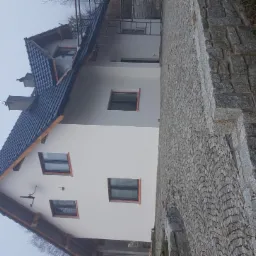 Świeżo otynkowany dom jednorodzinny z ciemną dachówką, widok z boku na elewację i kamienny chodnik.